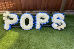 Blue & white funeral letters spelling pops. Completed with blue ribboned edging & white rose sprays